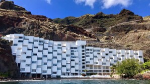 A modern white building with a unique modular design, situated by a body of water with rocky hills in the background.