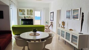 A modern living room with a green sofa, a dining table with chairs, a TV, a balcony with a view of the sea, and a white cabinet with glass doors.