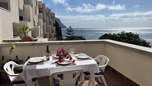 A dining table set on a balcony with a view of the sea, white chairs, and a bottle of wine.