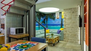A room with a kitchen area, a dining table with a colorful placemat, a sofa with cushions, a small coffee table, and a mural of a beach scene. - Petra design apartment Dubrovnik; FREE wireless internet and breakfast! (Dubrovnik)
