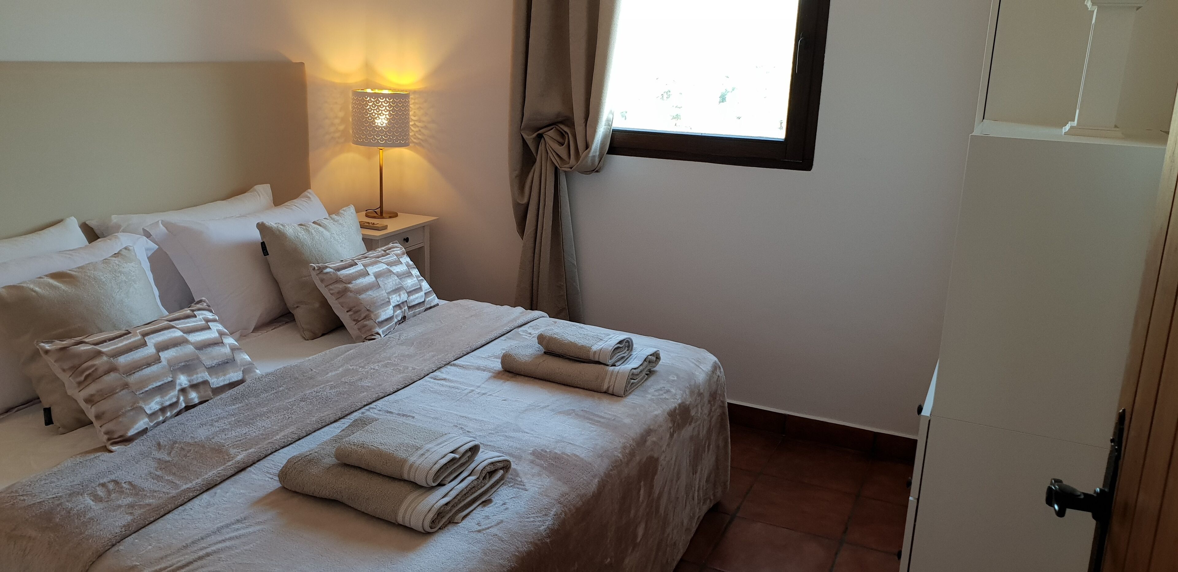 Una cama bien hecha con almohadas y toallas dobladas, una lámpara de noche y una ventana con cortinas.