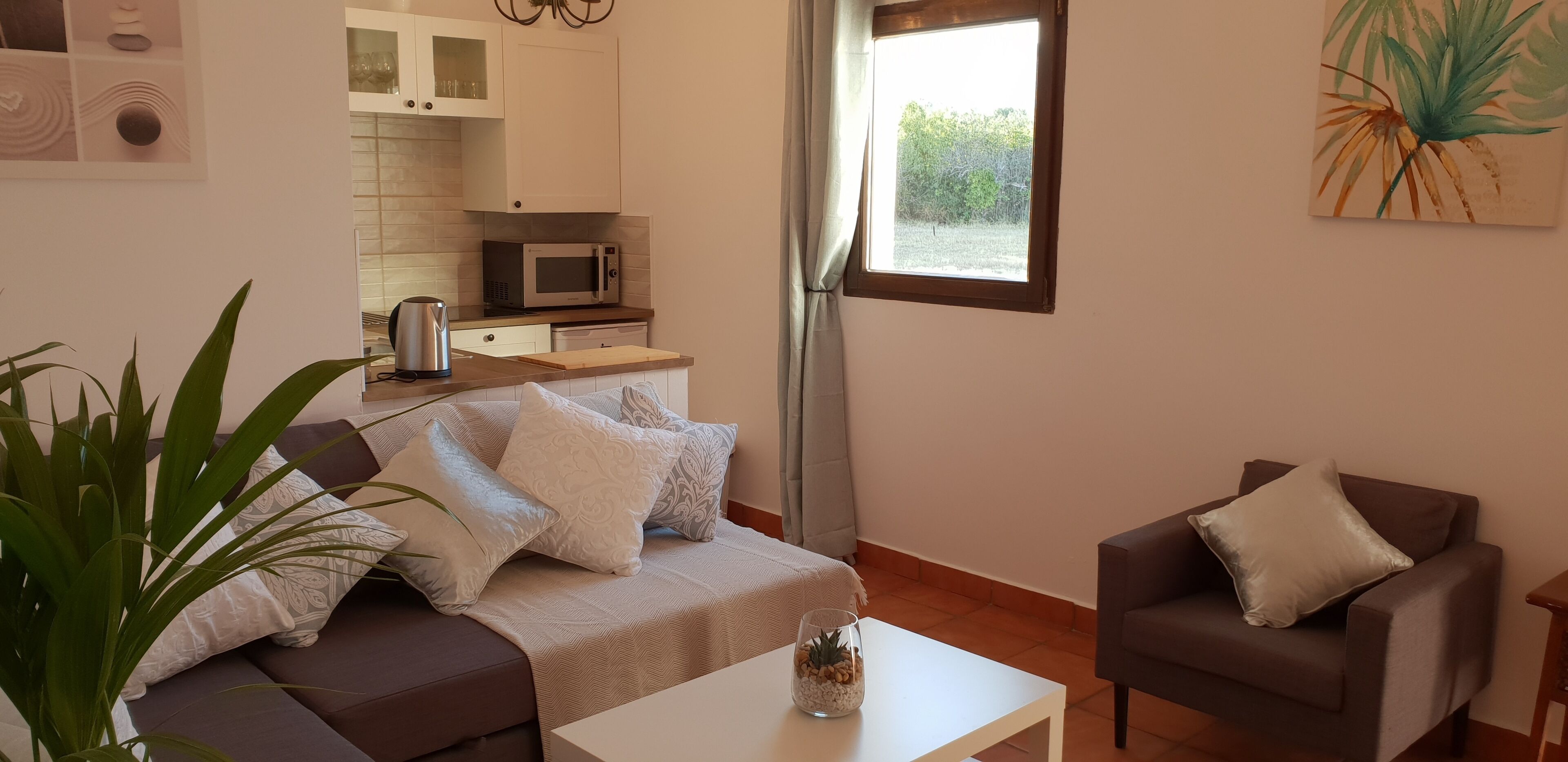 A living room with a sofa, a coffee table, and a side table. There is a kitchen area with a microwave and cabinets. A window with a view of greenery is visible.