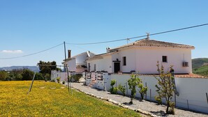 A two-story house with a white exterior and a tiled roof, surrounded by a garden with yellow flowers and a gravel path. - Cortijo el Chenil rural retreat, La Perrita (Casarabonela)