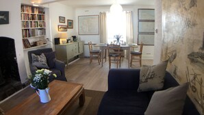 A living room with a sofa, a coffee table, a bookshelf, a dining area with a table and chairs, and a wall with artwork. - TRINITY COTTAGE. Charming historic cottage just off the Quay in Appledore. (Appledore)