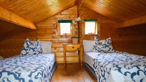 A cozy attic bedroom with wooden walls and a sloped ceiling. There are two beds with matching blue and white patterned bedding, a small wooden nightstand with a vase of flowers, and two windows with green curtains. - Los Palmeros cabin, ocean and mountain view (Vallehermoso)