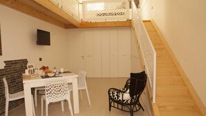 A dining area with a white table and chairs, a black chair with a patterned cushion, a brick wall, and a wooden staircase.