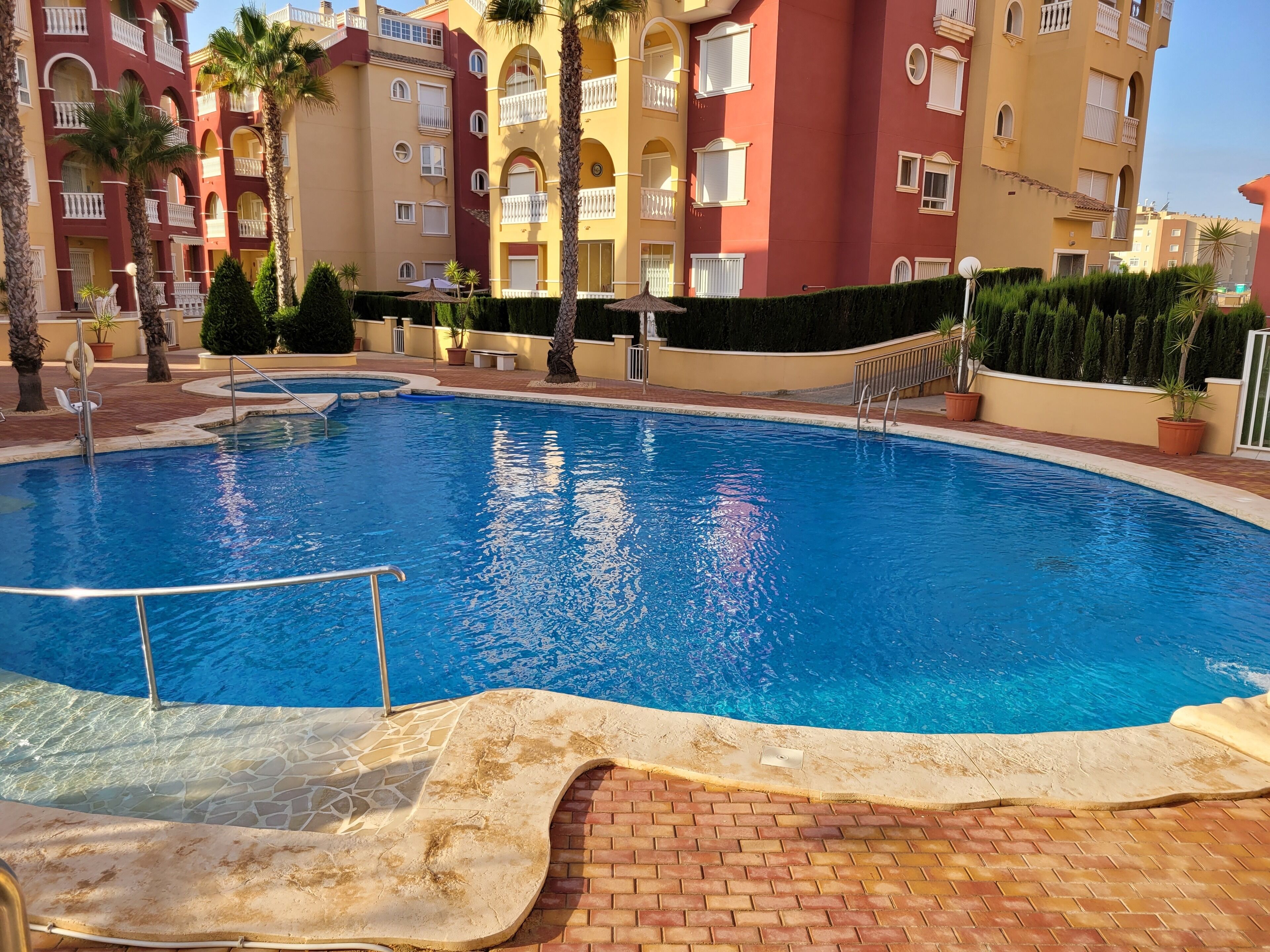 A swimming pool with a tiled deck, surrounded by palm trees and a multi-story building with balconies.