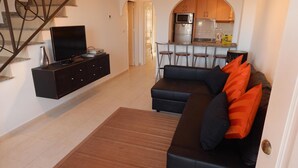 Una sala de estar con un sofá negro, un televisor y una cocina al fondo.