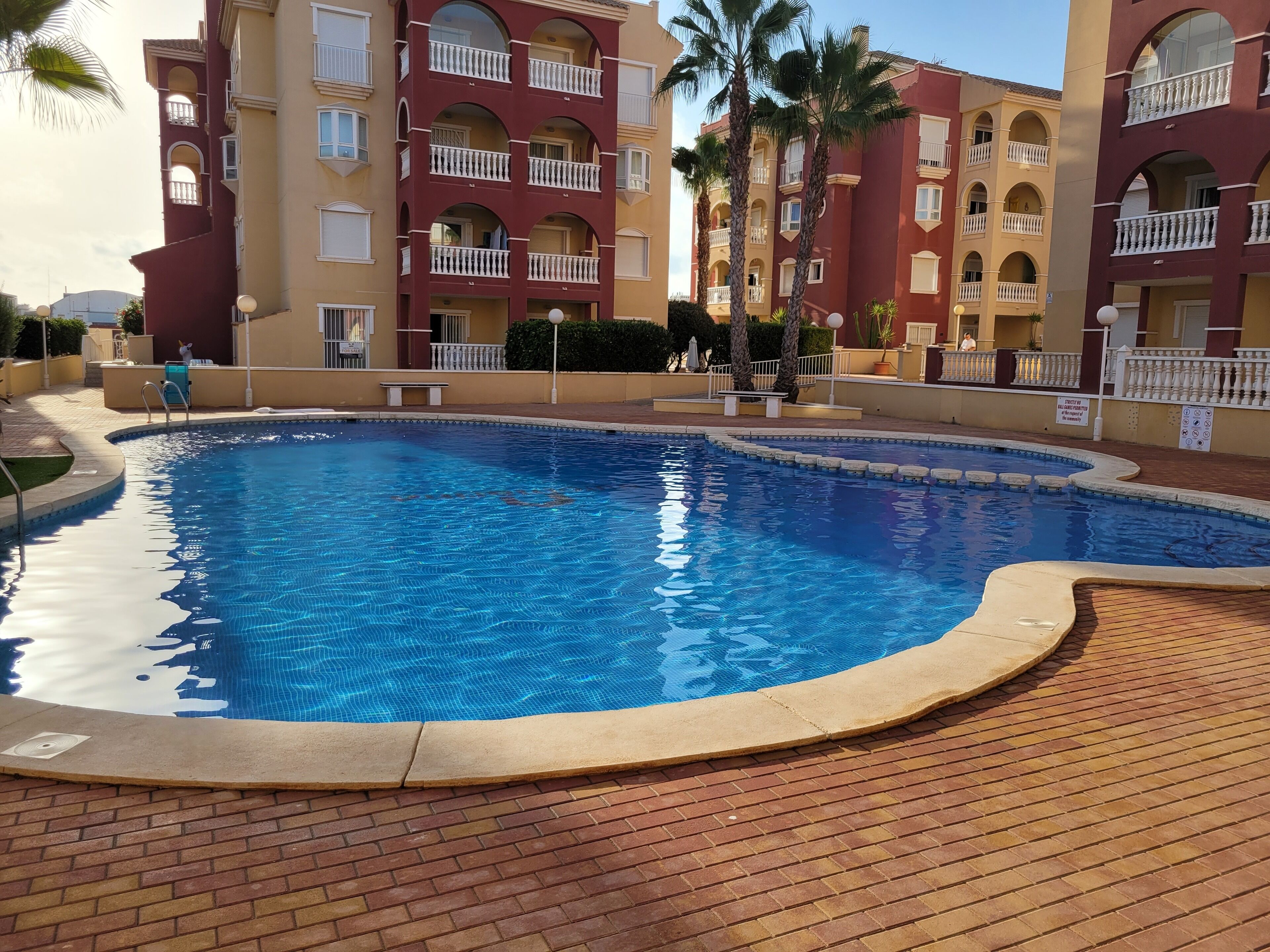Une piscine entourée d’une terrasse en briques, elle-même bordée d’immeubles à plusieurs étages dotés de balcons et de palmiers.