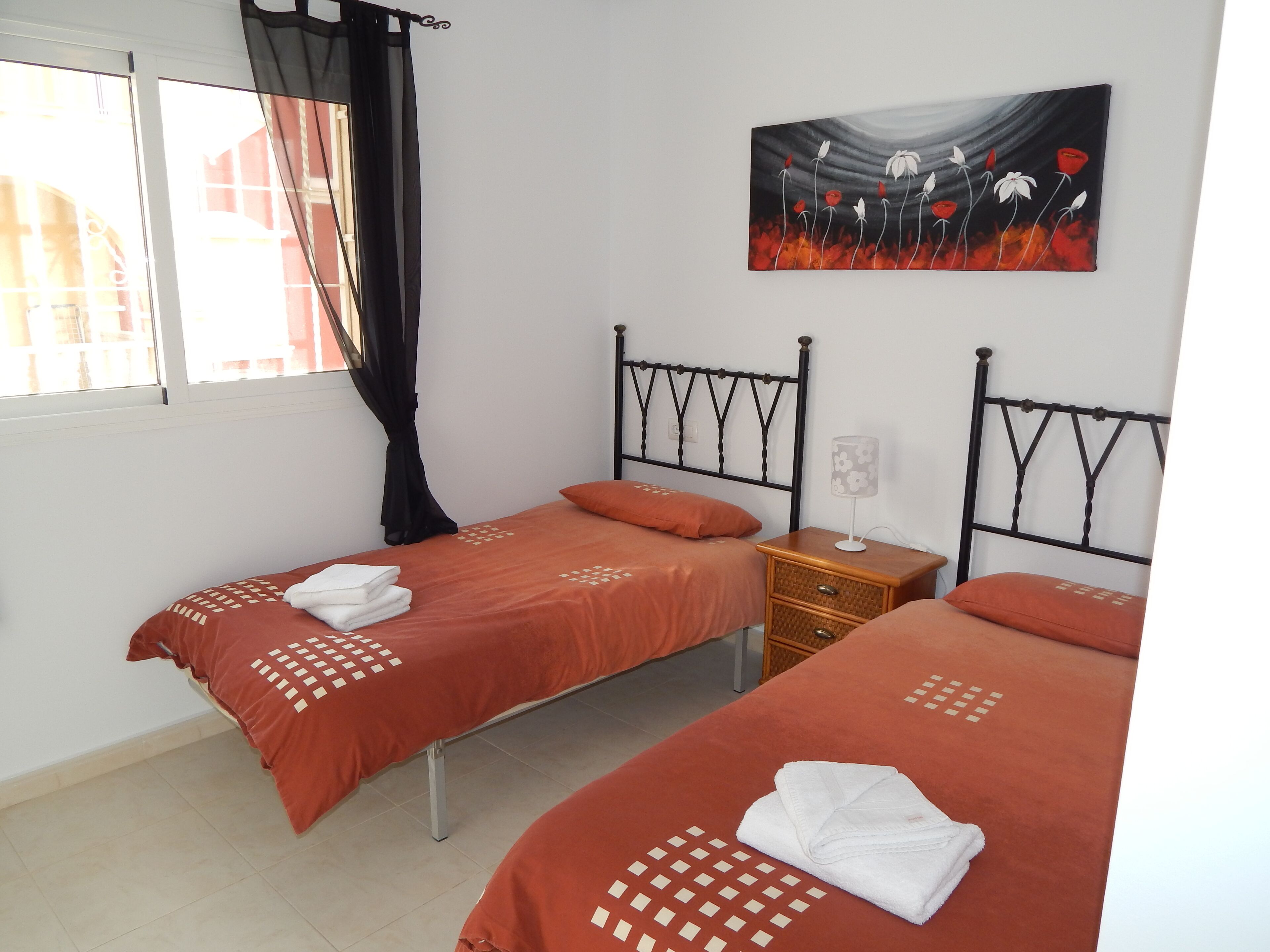 Ein Zimmer mit zwei Betten, jeweils mit roter Bettdecke und weißen Handtüchern. An der Wand hängt ein Gemälde, ein Nachttisch mit Lampe und ein Fenster mit Vorhängen.