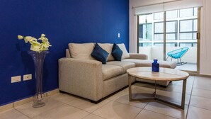 A living room with a beige sofa, a round coffee table, a blue vase with flowers, and a blue chair by a large window. - Central Huge PH with Private Rooftop and Jacuzzi (Playa del Carmen)