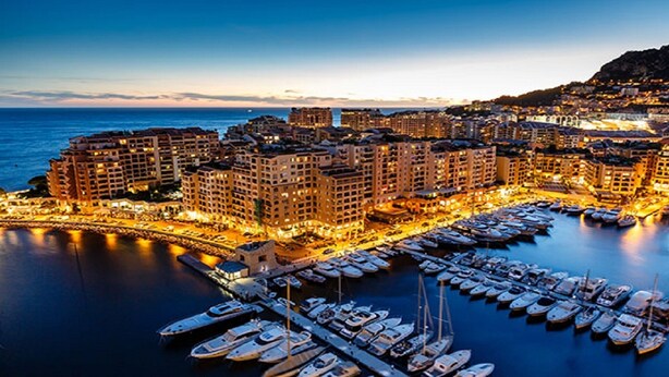 Un port de plaisance où de nombreux bateaux sont amarrés, entouré d’immeubles de grande hauteur dont les lumières diffusent une ambiance chaleureuse.