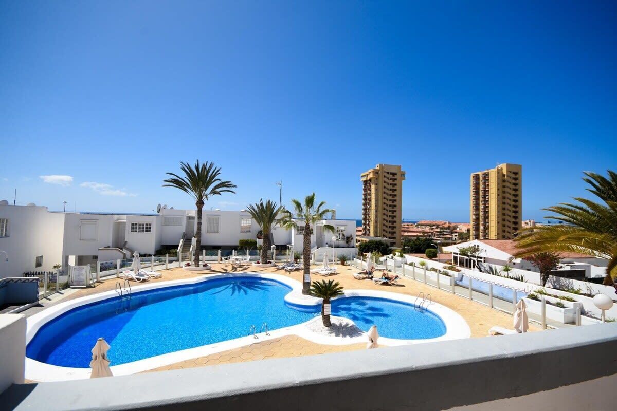 A swimming pool surrounded by palm trees and lounge chairs, with tall buildings in the background.