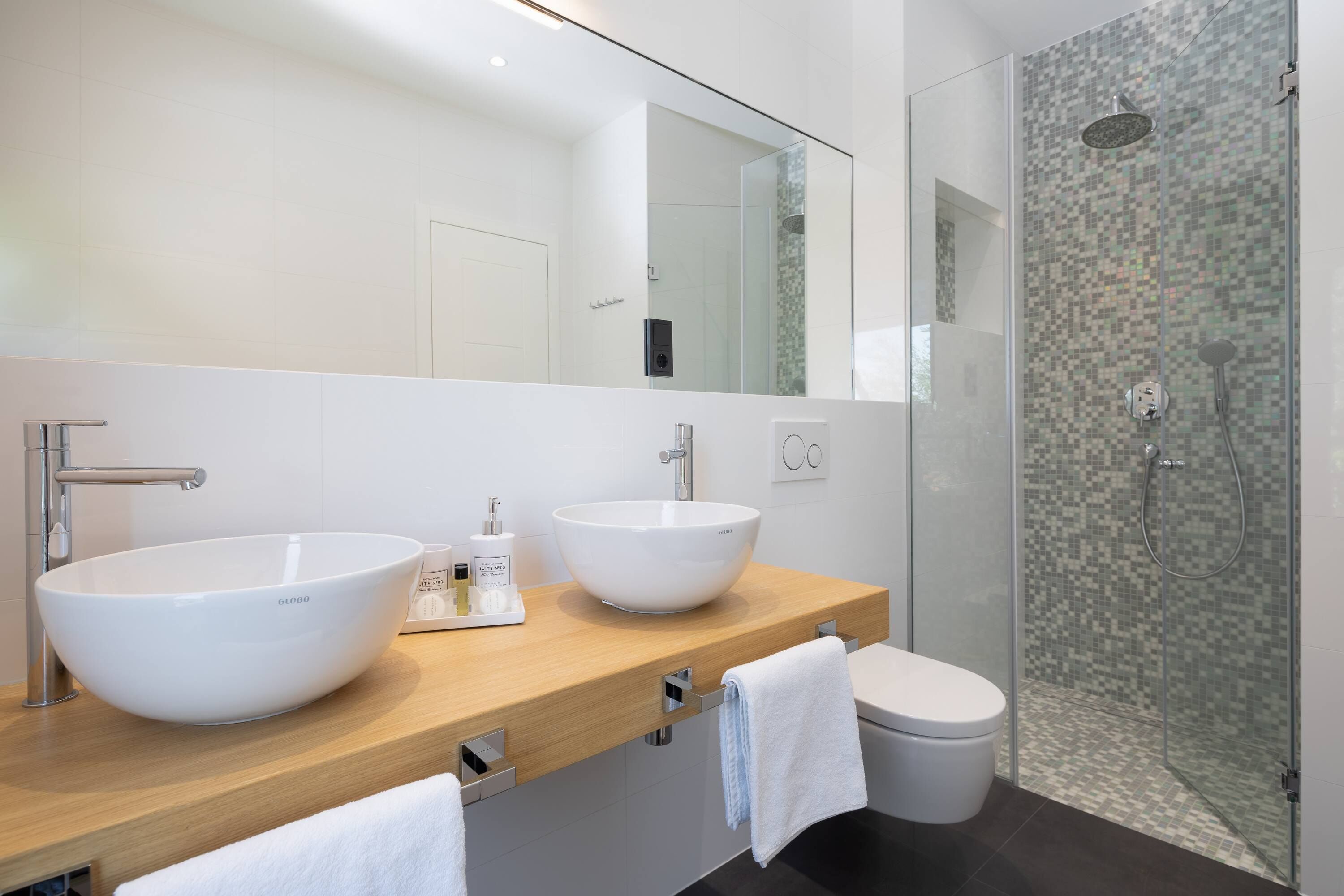 A modern bathroom with two large white vessel sinks, a wooden countertop, a glass-enclosed shower, and a white toilet.