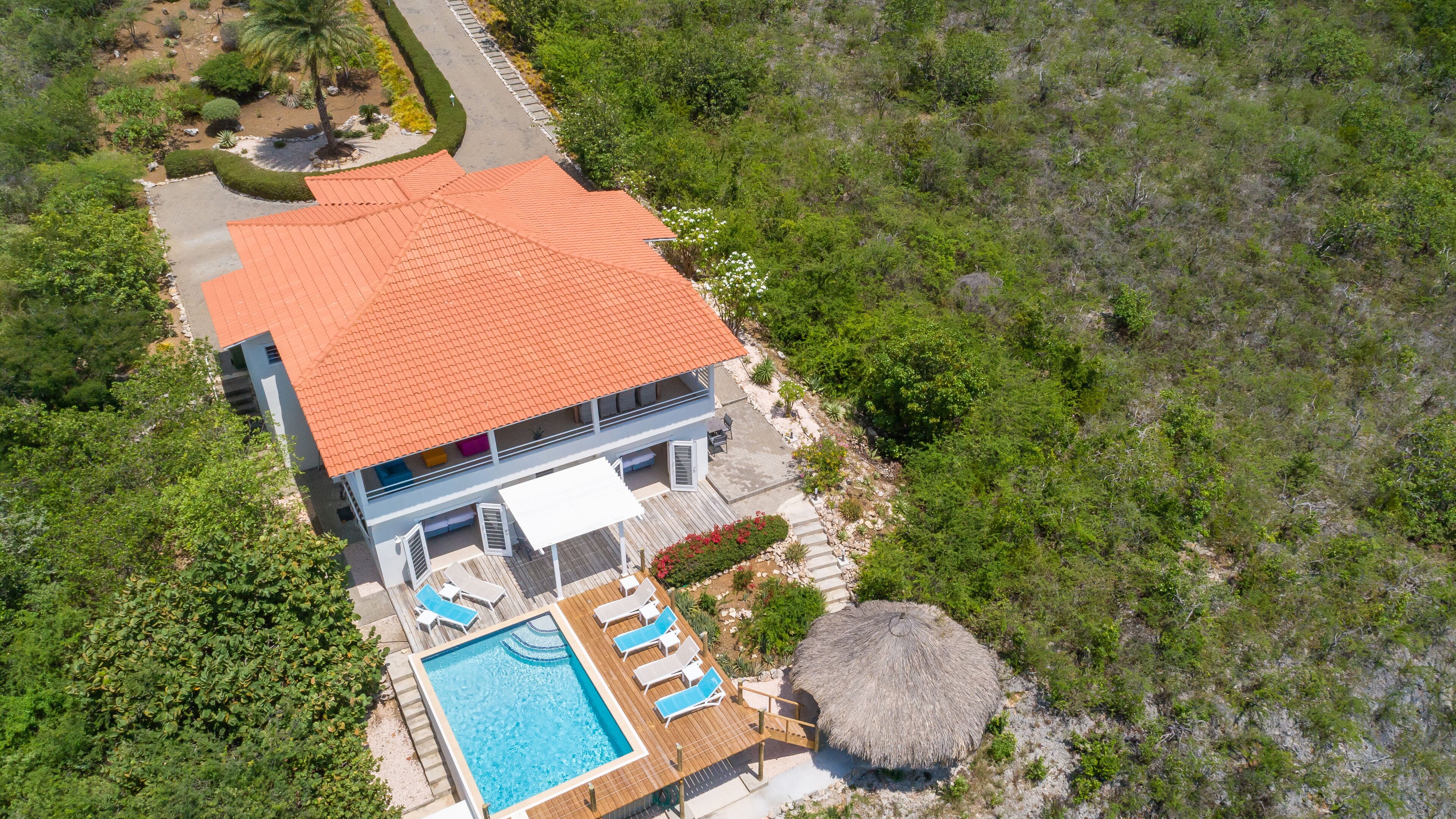 Vista aérea de una casa de dos pisos con techo de tejas rojas, piscina y una estructura con techo de paja.
