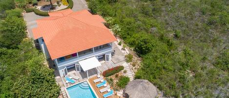 Vista aérea de una casa de dos pisos con techo de tejas rojas, piscina y una estructura con techo de paja.