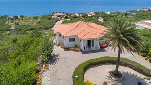 A coastal house with a red-tiled roof, surrounded by lush greenery and a palm tree. - Luxury Villa "Lions Cliff" with fantastic views over the Caribbean Sea (St. Willibrordus)