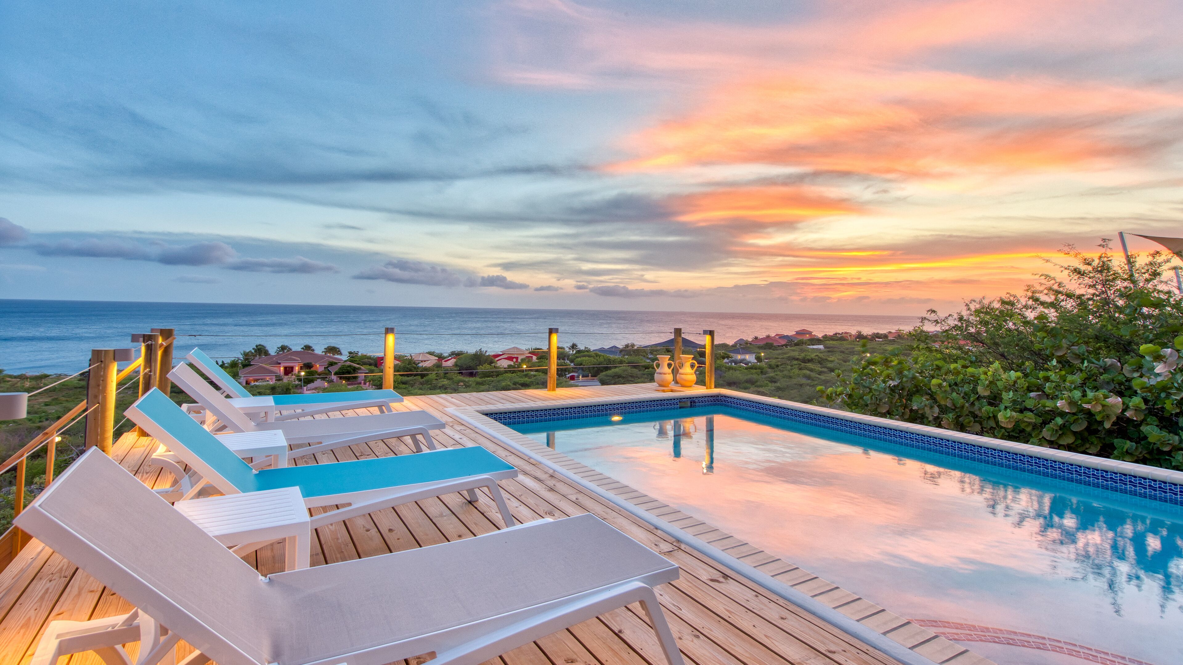 Área da piscina com espreguiçadeiras, deck de madeira e vista para o oceano ao pôr do sol.
