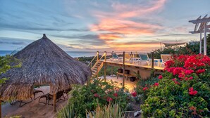 A beachside resort with a thatched-roof hut, a wooden deck with lounge chairs, and a view of the sunset over the ocean. - Luxury Villa "Lions Cliff" with fantastic views over the Caribbean Sea (St. Willibrordus)