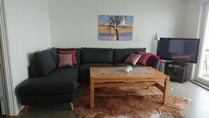 Sala de estar com um sofá preto, mesa de centro de madeira, televisão e um quadro de uma árvore.