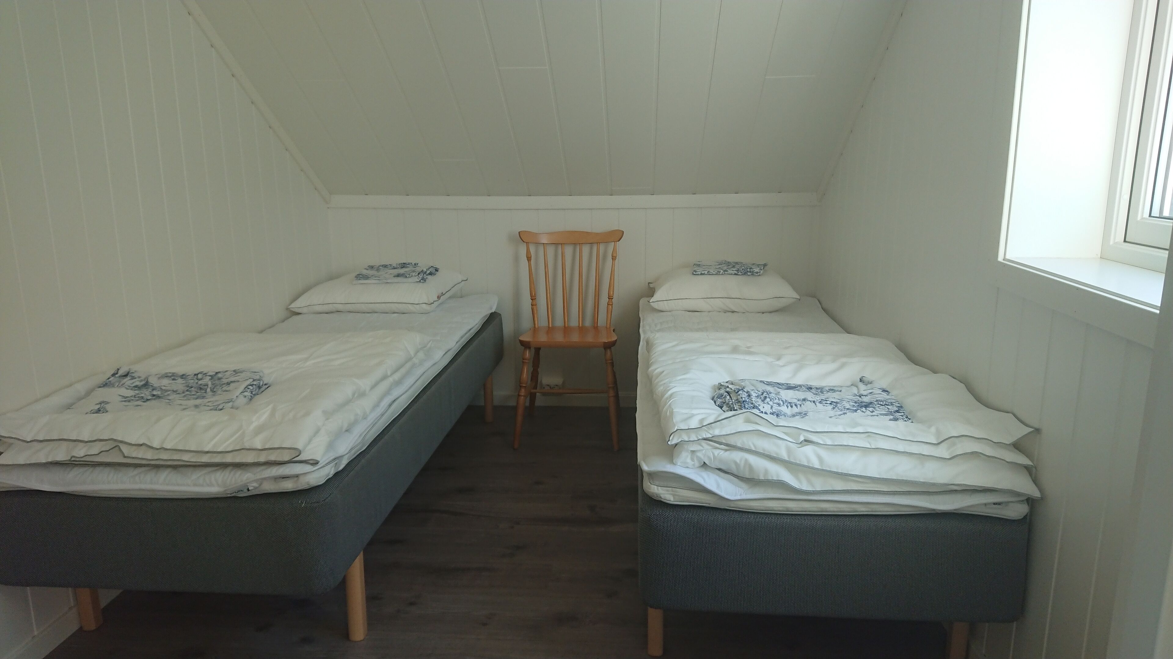 Une petite pièce faiblement éclairée, avec deux lits simples, une chaise en bois et une fenêtre.