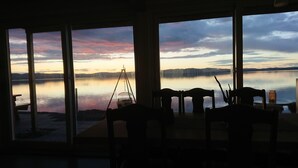 Quarto com grandes janelas dando para um lago calmo ao pôr do sol, com um tripé e cadeiras.