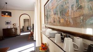 Un área de comedor con un frutero, platos blancos y tazas de café sobre una encimera. Un cuadro enmarcado de una escena histórica adorna la pared.