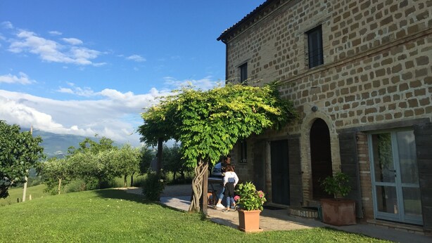 Un edificio in pietra con un portico coperto, un prato e un albero con verde chioma.