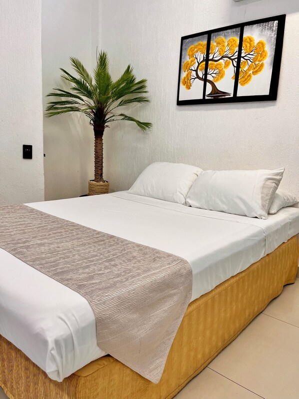 A bed with a white bedspread and a patterned throw, a single white pillow, a small potted plant, and a framed picture of a tree on the wall.