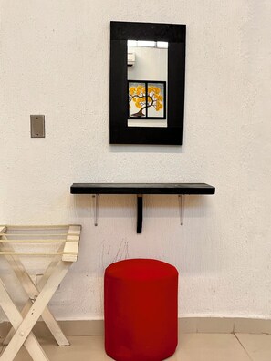 A room with a white textured wall, a black-framed mirror, a wooden stool, and a red cylindrical object.