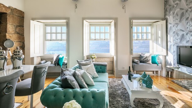 A spacious living room with a teal sofa, two windows with views, a stone wall, and a flat-screen TV. - Oporto Platinum Loft // Luxury Accommodation// Unforgettable Douro River View. (Porto)