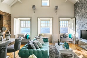 A spacious living room with a teal sofa, two windows with views, a stone wall, and a flat-screen TV. - Oporto Platinum Loft // Luxury Accommodation// Unforgettable Douro River View. (Porto)