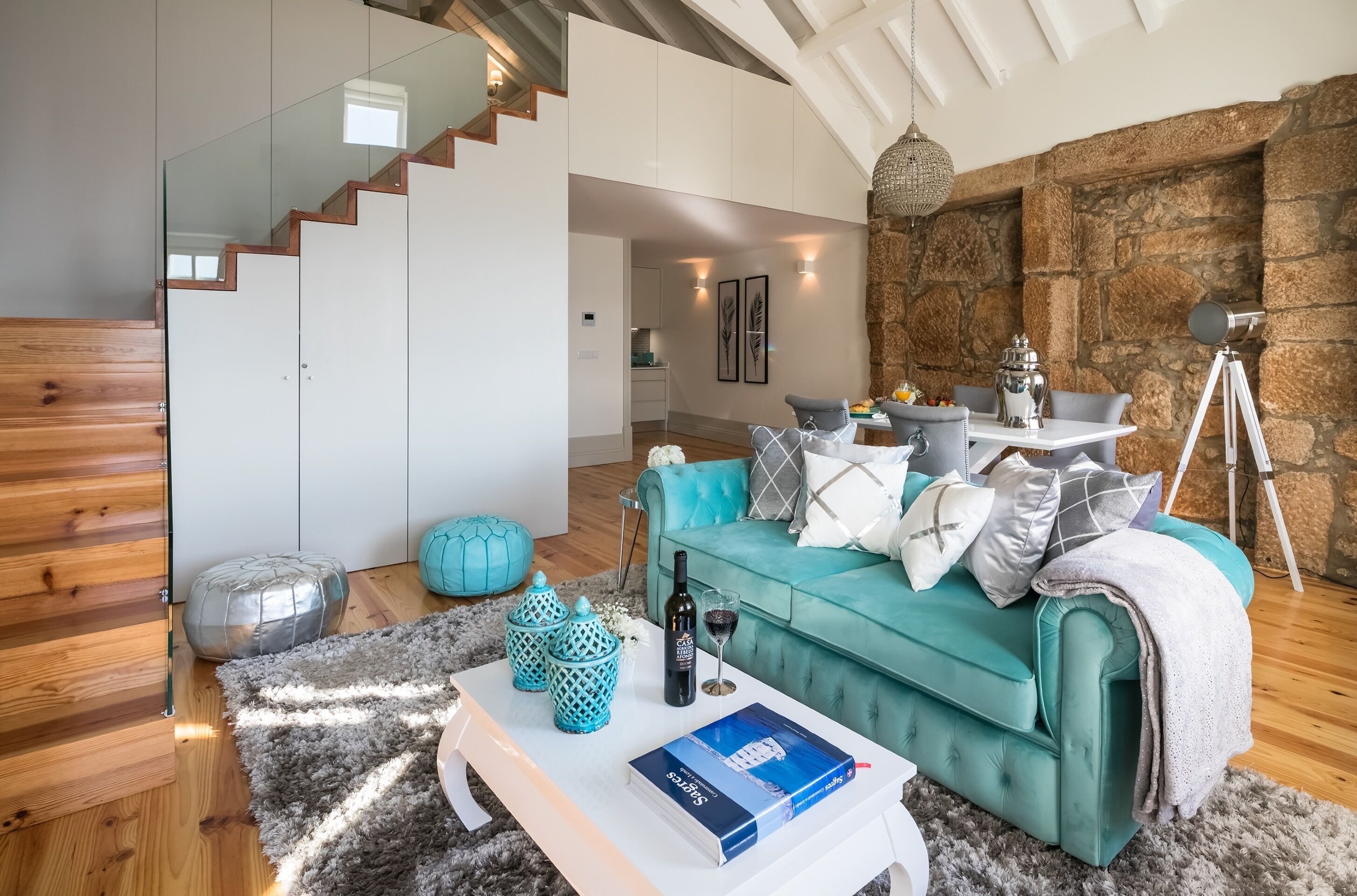 A modern living room with a teal sofa, a white coffee table, and a staircase with glass railings.