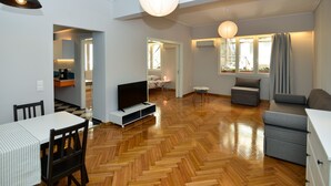 Ein modernes Wohnzimmer mit Holzdielenmuster, einem Flachbildfernseher, einem grauen Sofa und einem Essbereich mit weißem Tisch und Stühlen.