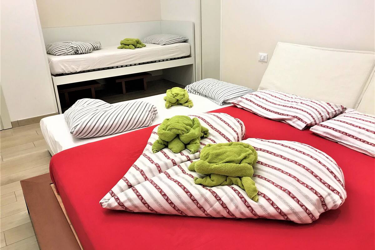 A hotel room with a red bedspread, striped pillows, and a bunk bed in the background.