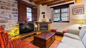 Ein gemütliches Wohnzimmer mit Steinwand, Holzbalken, Kamin, Fernseher und einer Couch.