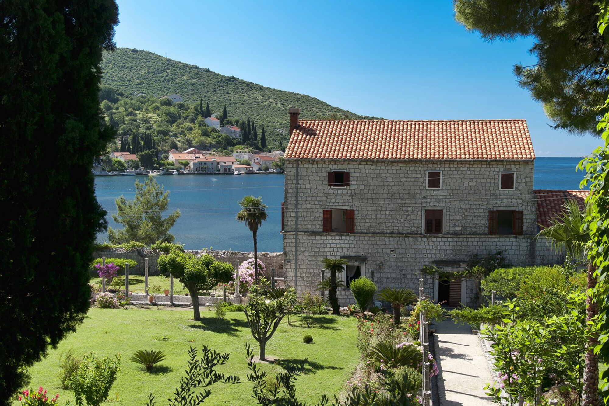 A stone building with a tiled roof, surrounded by a well-maintained garden and a view of a body of water and hills in the background.