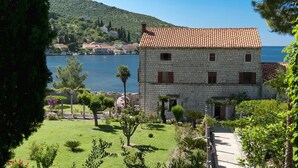 A stone building with a tiled roof, surrounded by a well-maintained garden and a view of a body of water and hills in the background. - Room 5 a/c park view Beach&Sea front Castle parking (Zaton)