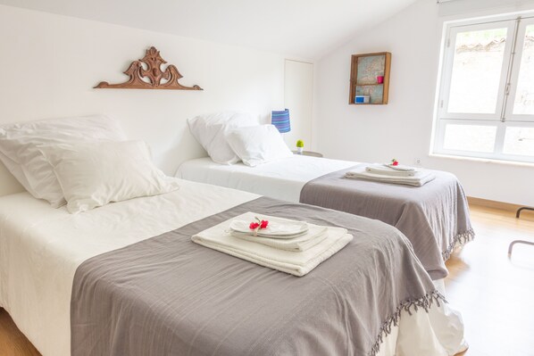 A hotel room with two beds, each with white linens and a grey blanket. There is a wooden headboard, a small table with a lamp, and a window with a view of the outside.