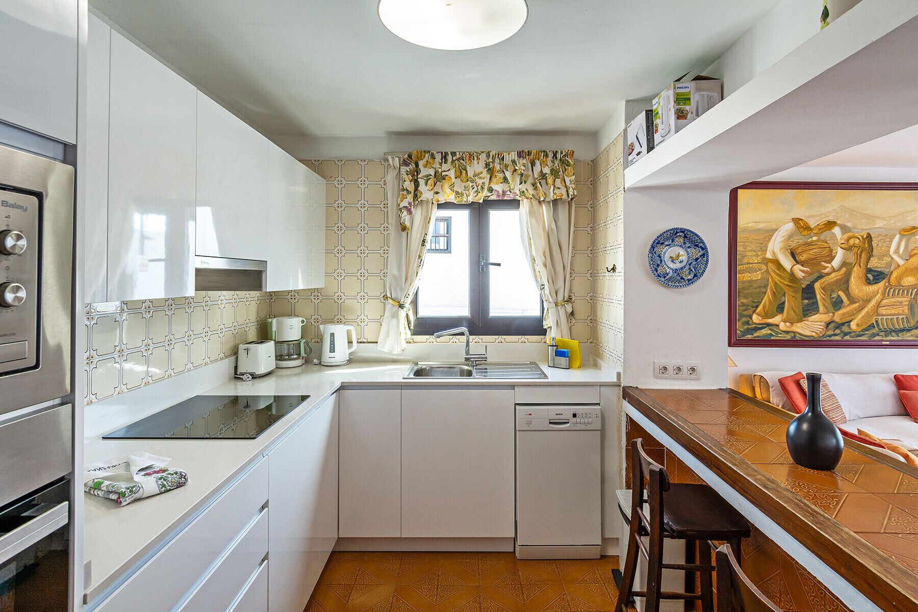 Une cuisine compacte équipée d’un lave-vaisselle, d’une cuisinière et d’un évier. Il y a une fenêtre avec des rideaux et un tableau accroché au mur.