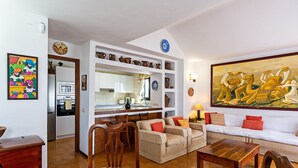 Un salon comprenant un canapé, des fauteuils, une table basse et un coin cuisine avec des étagères.