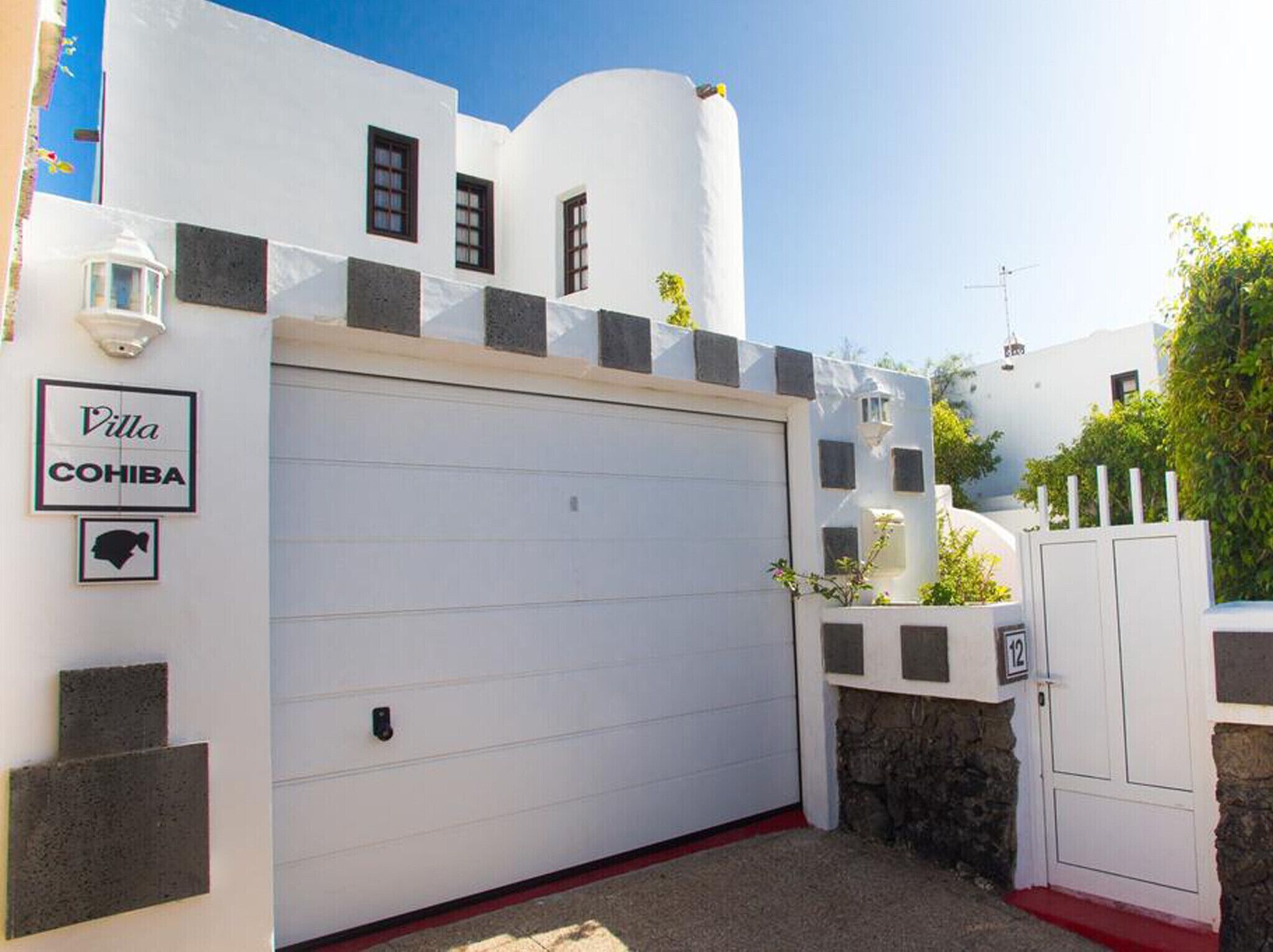 Una villa bianca con un portone garage, un cartello con scritto "Villa COHIBA" e un piccolo giardino con un cancello con il numero 12.