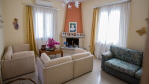 Un salon avec des canapés de couleur beige, un fauteuil à motifs, une cheminée ornée de photos encadrées et un lustre.