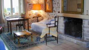 A cozy room with a stone wall, a bed with a white bedspread, a wooden chair, a small table, and a fireplace. - Chevannes: House with character (Chevannes)