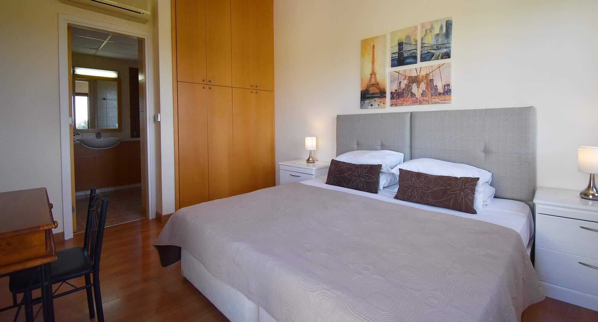 A hotel room with a large bed, a desk, a chair, a wardrobe, and artwork on the wall.