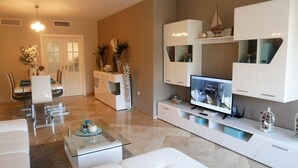 Un salón moderno con una mesa de centro de vidrio, sofás blancos, un televisor de pantalla plana y un comedor con una mesa y sillas blancas.