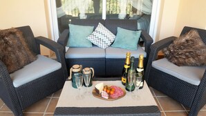 Un patio amueblado con muebles de mimbre, un sofá con cojines y una mesa dispuesta con botellas de vino, copas y una tabla de embutidos.