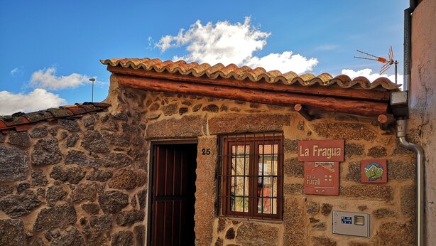 A rustic stone building with a tiled roof, a wooden door, and a window with bars. There is a sign on the wall that reads "La Fragua casa rural" and a number "25" above the door.