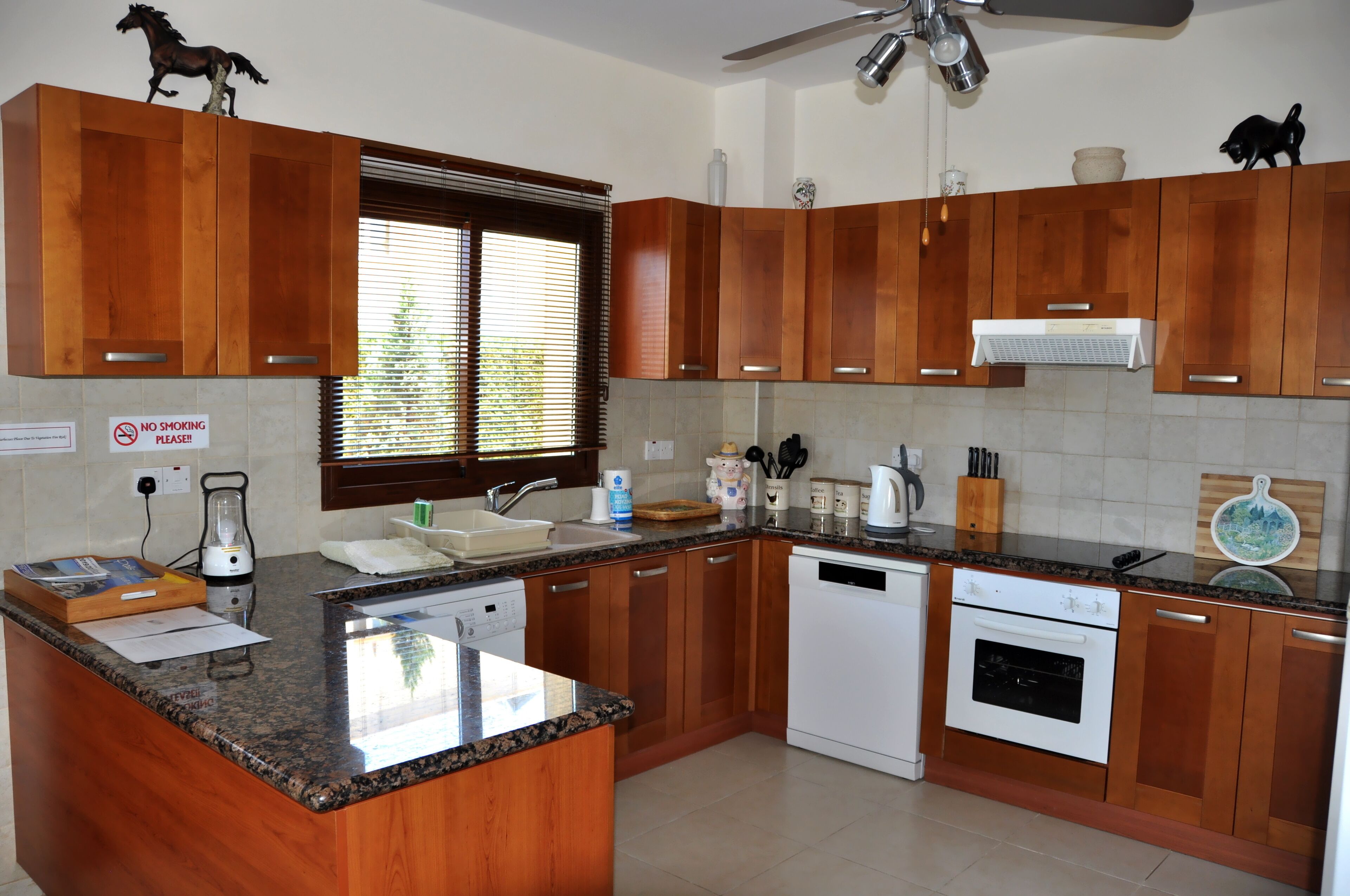 Une cuisine avec des meubles en bois, un plan de travail en granit, une cuisinière et une fenêtre avec des stores.