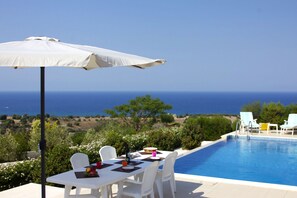 Un espace repas aménagé au bord de la piscine, avec une table dressée pour deux personnes, un parasol blanc et une vue sur la mer.